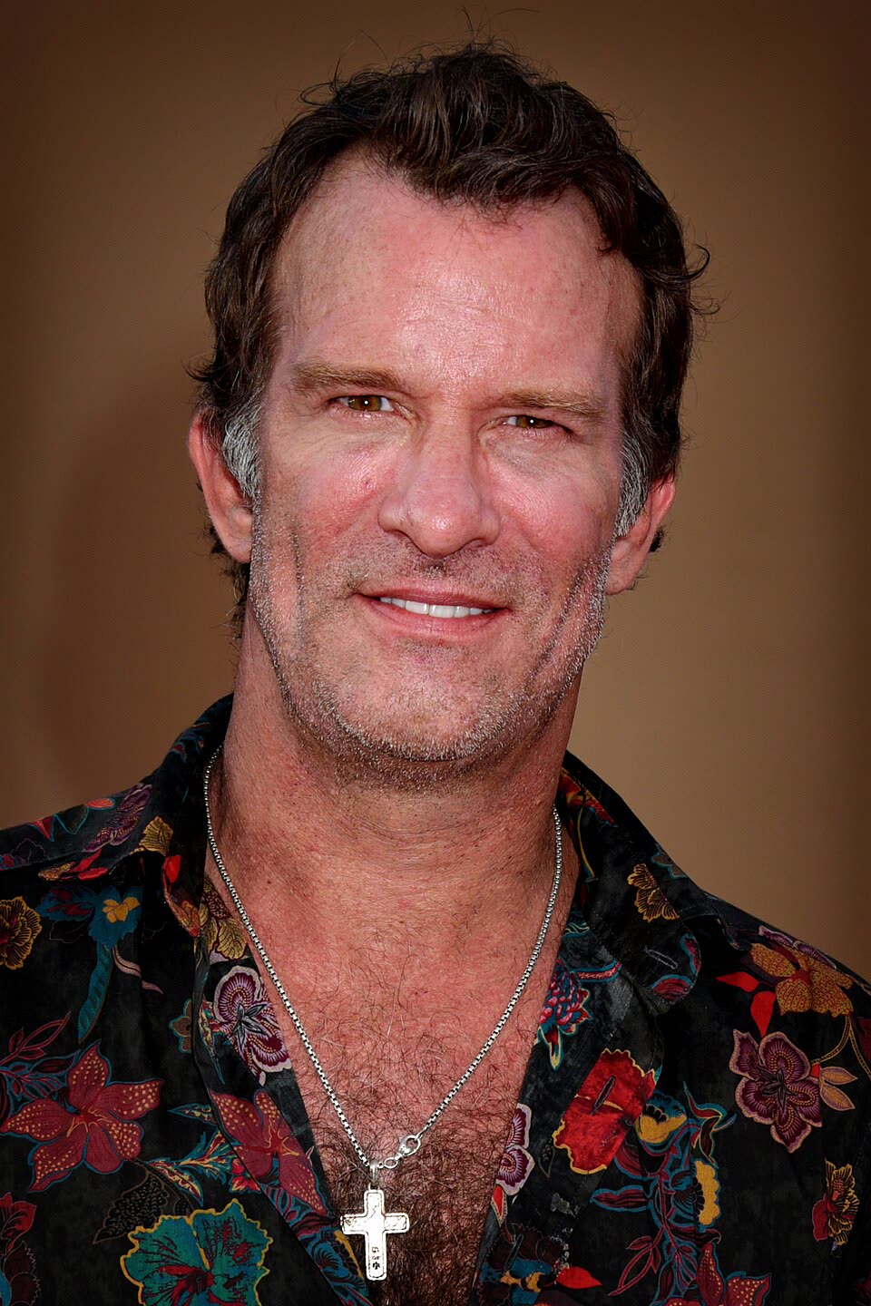 Portrait of actor Thomas Jane smiling, wearing a dark floral shirt and a silver cross on a chain.