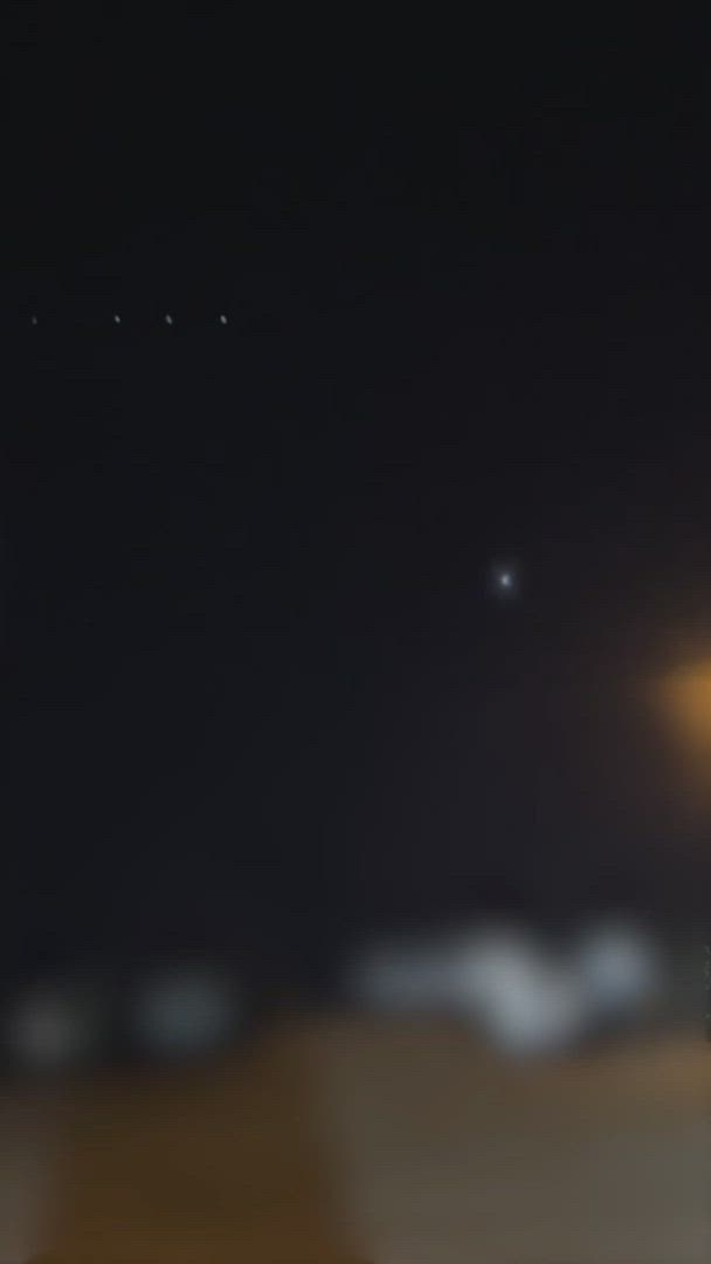 A dark vertical cellphone video frame at night showing a single point of light above a dim horizon.