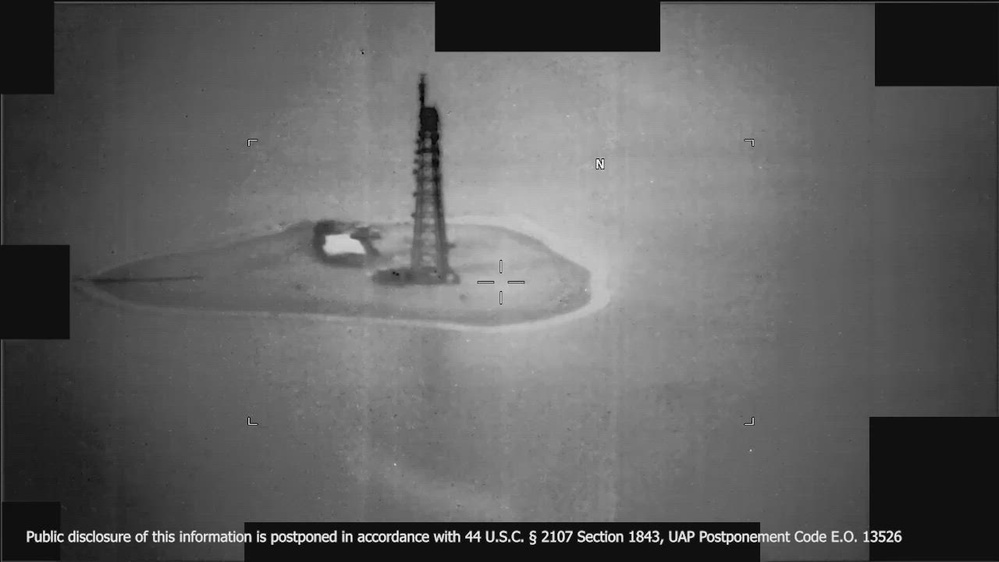 Black-and-white infrared frame of a small low island with a tall lattice tower, surrounded by water.
